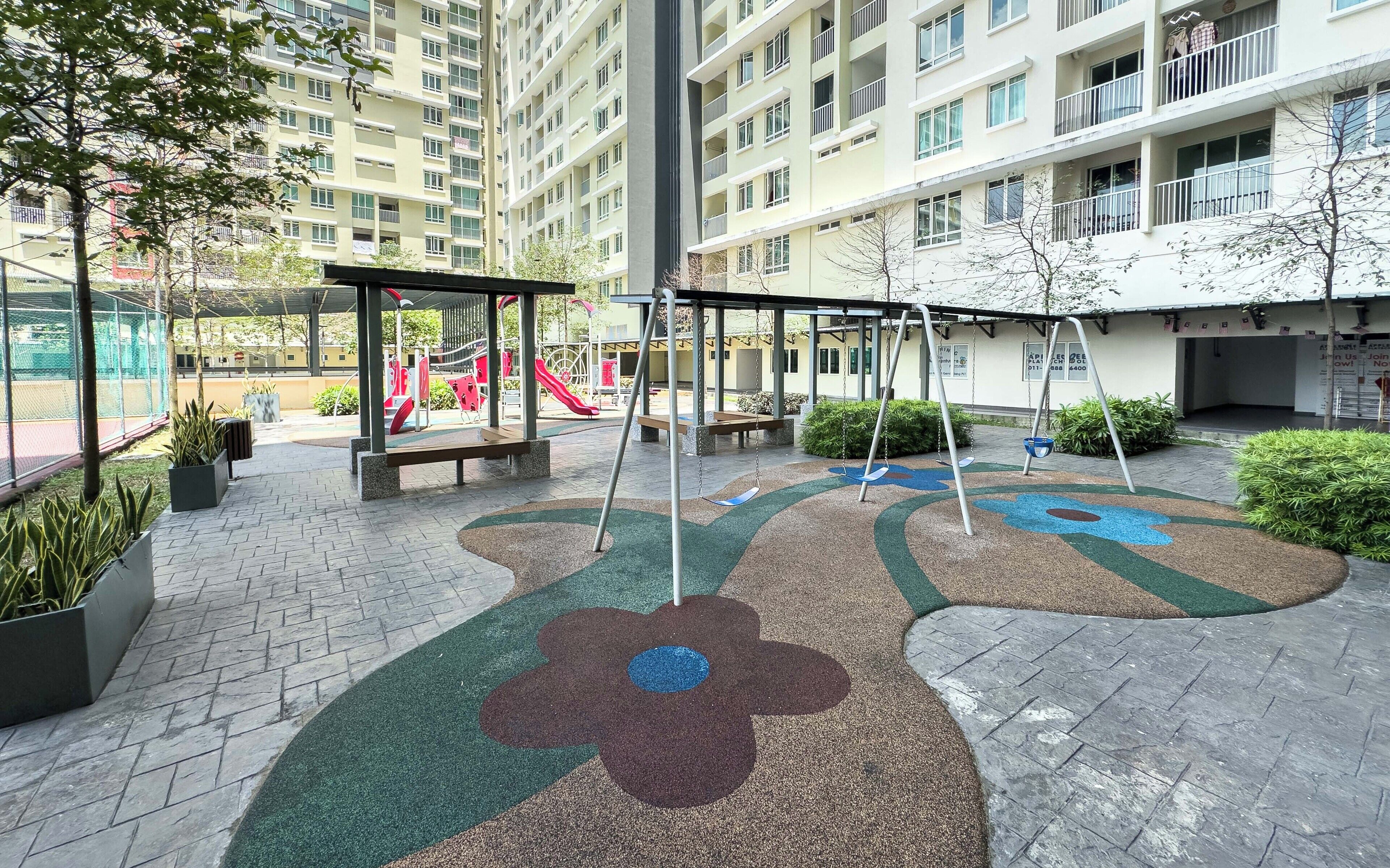 A playground with swings and a slide, surrounded by a paved area with planters and a building in the background.