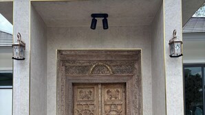 Eine große, kunstvoll verzierte Holztür mit feinen Schnitzereien, begleitet von zwei laternenartigen Leuchten, in ein modernes Gebäude mit klarem, minimalistischem Design eingesetzt.