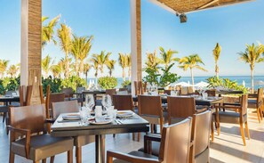 An outdoor dining area with tables set for two, overlooking a beach with palm trees and the ocean.