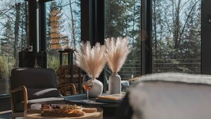 A cozy room with a large window, a fireplace, and a round table with food and drinks. - Ökohaus Mont-Tremblant: Private Luxury Nordic Eco Cabin with Spa & Sauna (La Conception)