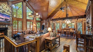 A spacious lodge with a stone fireplace, wooden furnishings, and large windows offering views of the surrounding forest. - SUNNYSIDE, FLAT YARD, HOT TUB, MOUNTAIN VIEWS, WOOD BURNING FIREPLACE (Sundance)