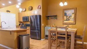 A modern kitchen with wooden cabinets, a stainless steel refrigerator, a microwave, and a dining area with a wooden table and chairs. - Cozy 2-Bedroom Condo with Mountain View, Hot tub and Heated Pools (Mesa)