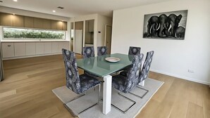 Ett modernt matområde med ett glasbord och mönstrade stolar, ett kök med utsikt genom fönstret och ett elefantmotiv som hänger på väggen.