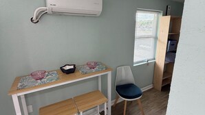 A room with a wooden desk, two benches, a white chair, a window with blinds, and an air conditioning unit mounted on the wall. - Beach Studio steps to the Beach (Cocoa Beach)