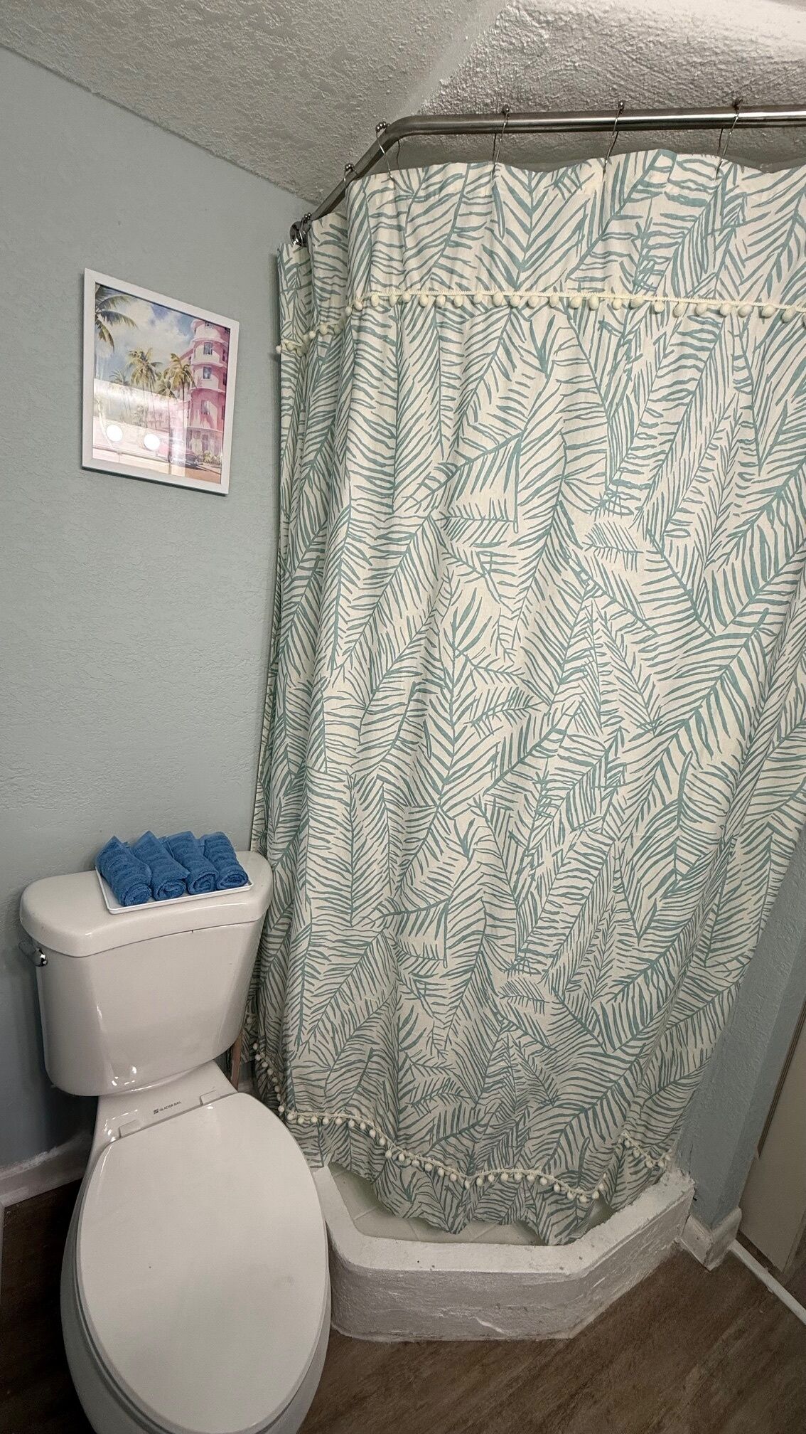 A bathroom with a toilet, a shower curtain with a leaf pattern, and a framed picture on the wall.