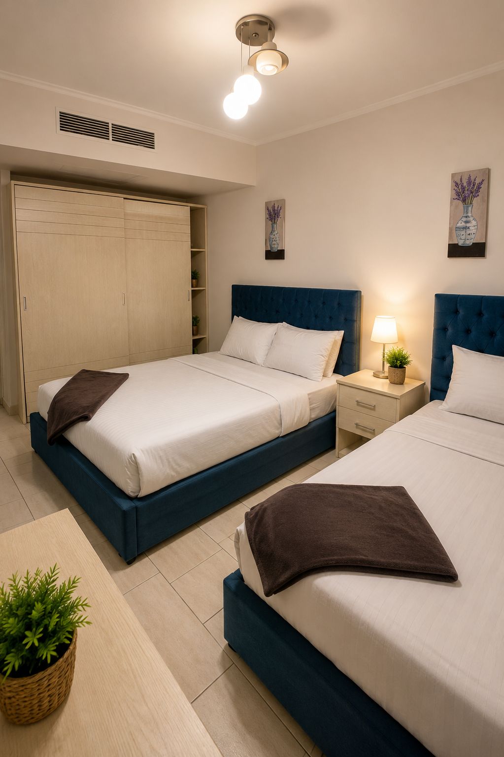 Una habitación de hotel con dos camas, un armario, una mesita de noche con lámpara y una planta pequeña.