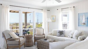 Ein helles, weißes Interieur mit Korbmöbeln, einem Deckenventilator und großen Fenstern mit Blick auf den Strand.