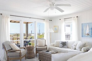 A bright, white interior with wicker furniture, a ceiling fan, and large windows offering a view of the beach. - Oceanfront Oasis + Guesthouse: 2 Hot Tubs, Pool! (Atlantic Beach)
