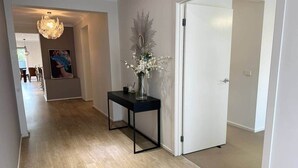 Käytävän lattia on puuta, ja siellä on musta metallinen konsolipöytä, jossa on maljakko täynnä kukkia, seinälle kiinnitetty peili ja valkoinen ovi.