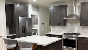 A modern kitchen with dark cabinets, a stainless steel refrigerator, an oven, and a marble countertop island with two bar stools. - Enjoyable 4-bedroom house with a beautiful rock water fall slide swimming pool. (Ontario)