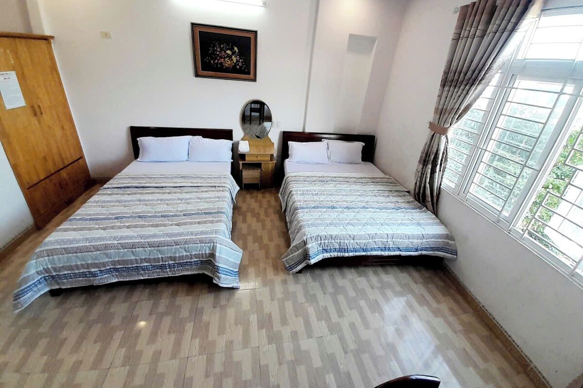 A room with two beds, a wooden wardrobe, a small table, a mirror, and a window with curtains.