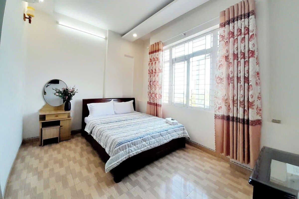A bedroom with a bed, a bedside table with a mirror, a small cabinet, and a window with curtains.
