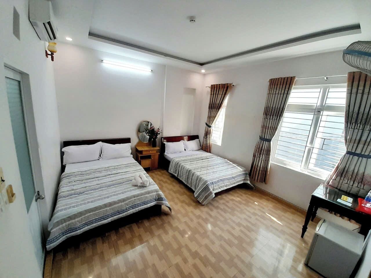 A hotel room with two beds, a wooden floor, a desk with a chair, a window with curtains, and a ceiling fan.