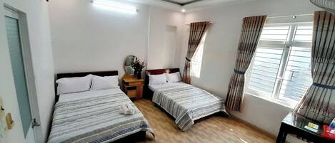 A hotel room with two beds, a wooden floor, a desk with a chair, a window with curtains, and a ceiling fan.