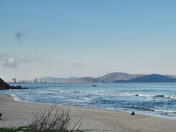 A beach with a clear view of the sea, distant mountains, and a city skyline in the background.
