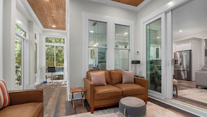A modern living room with a brown leather sofa, a round ottoman, and a glass door leading to a kitchen. - Enjoy sun-kissed Island days at this recently-built four-bedroom/ 3.5-bath (Bald Head Island)