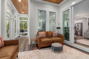 A modern living room with a brown leather sofa, a round ottoman, and a glass door leading to a kitchen. - Enjoy sun-kissed Island days at this recently-built four-bedroom/ 3.5-bath (Bald Head Island)