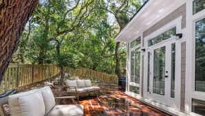 A wooden deck with outdoor furniture, surrounded by trees and a white building with large windows. - Enjoy sun-kissed Island days at this recently-built four-bedroom/ 3.5-bath (Bald Head Island)