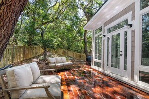 A wooden deck with outdoor furniture, surrounded by trees and a white building with large windows. - Enjoy sun-kissed Island days at this recently-built four-bedroom/ 3.5-bath (Bald Head Island)
