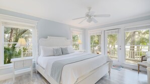 Ein geräumiges Schlafzimmer mit einem großen Bett, einem Deckenventilator und großen Fenstern mit Blick auf Palmen.