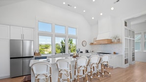 A modern kitchen with white cabinetry, a stainless steel refrigerator, a large island with wicker chairs, and a view of greenery outside. - VIEWS of the Atlantic will mesmerize you from The Glass Slipper (Bald Head Island)