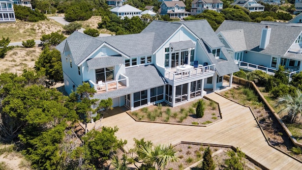 A large, light blue house with multiple levels and large windows, surrounded by greenery and other houses. - VIEWS of the Atlantic will mesmerize you from The Glass Slipper (Bald Head Island)