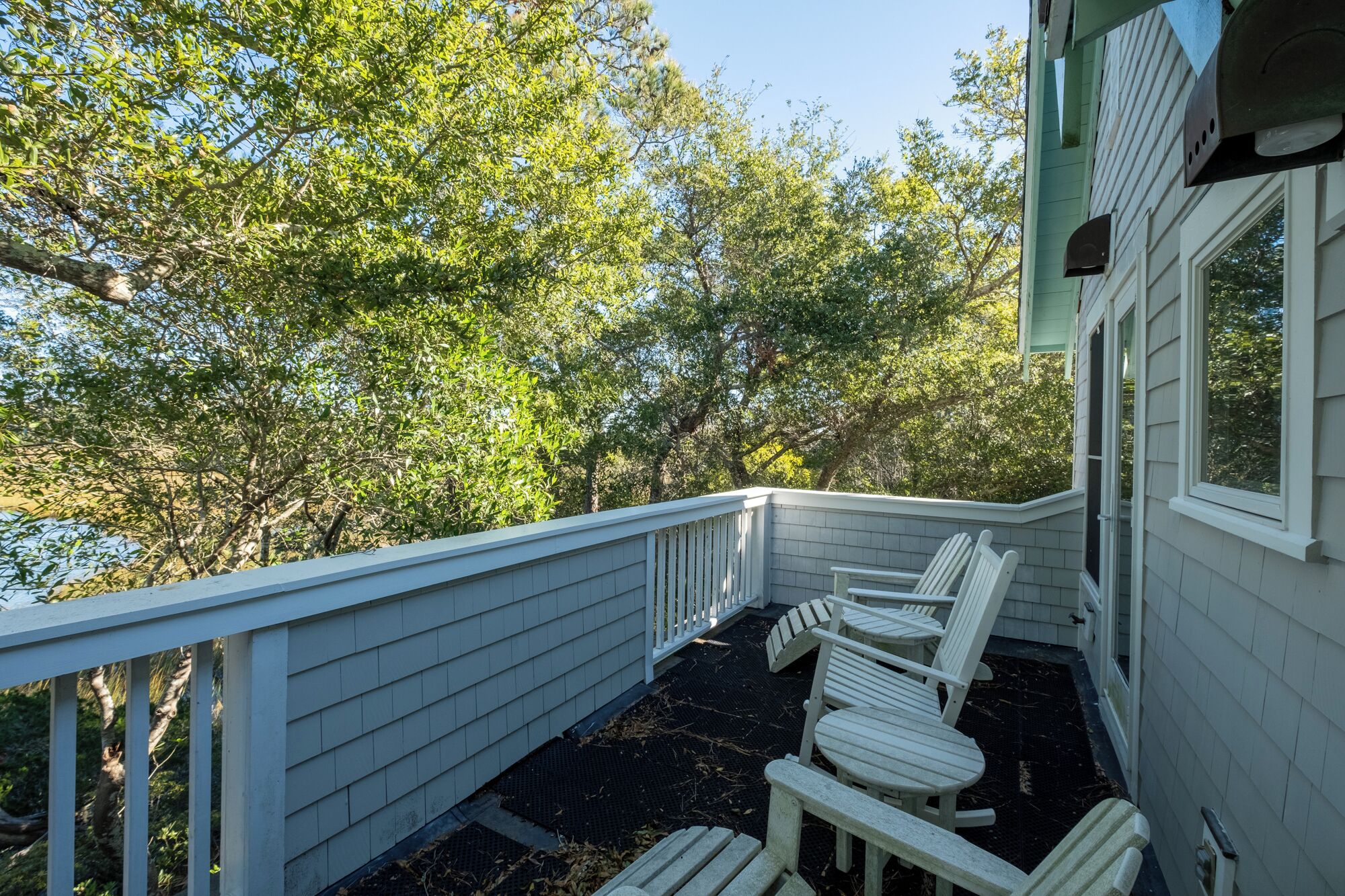 A covered outdoor area with a white railing, a set of Adirondack chairs, and a view of trees and a building.