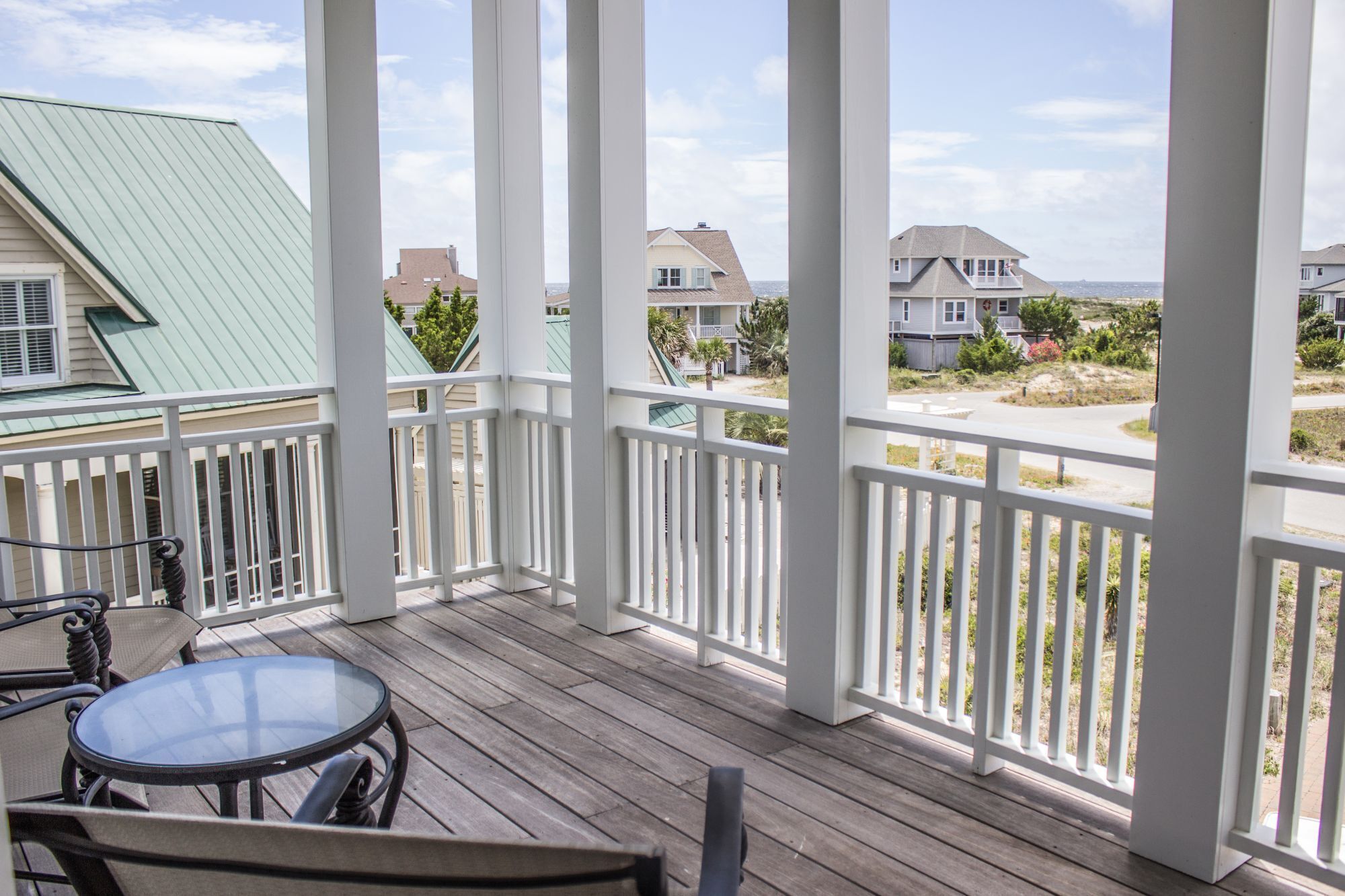 A wooden deck with a glass table and chairs, overlooking a residential area with houses and a beach.