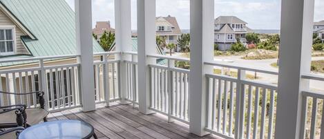 A wooden deck with a glass table and chairs, overlooking a residential area with houses and a beach.
