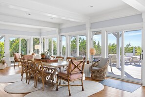 A spacious dining area with a large table and multiple chairs, large windows offering views of greenery, and a deck area with outdoor furniture. - Poolside Paradise is luxury vacationing! Enjoy beautiful sunsets (Bald Head Island)