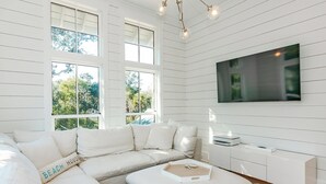 A modern living room with a sectional sofa, a flat-screen TV, and a large window with a view of trees. - Your dream vacation retreat is nestled in the Maritime Forest (Bald Head Island)