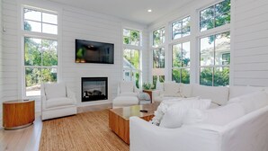 A spacious living room with a fireplace, white furniture, large windows, and a flat-screen TV. - Your dream vacation retreat is nestled in the Maritime Forest (Bald Head Island)