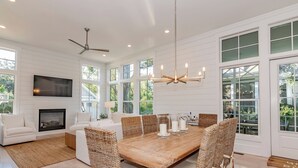 A spacious living room with a fireplace, a large dining table with wicker chairs, and a ceiling fan. - Your dream vacation retreat is nestled in the Maritime Forest (Bald Head Island)