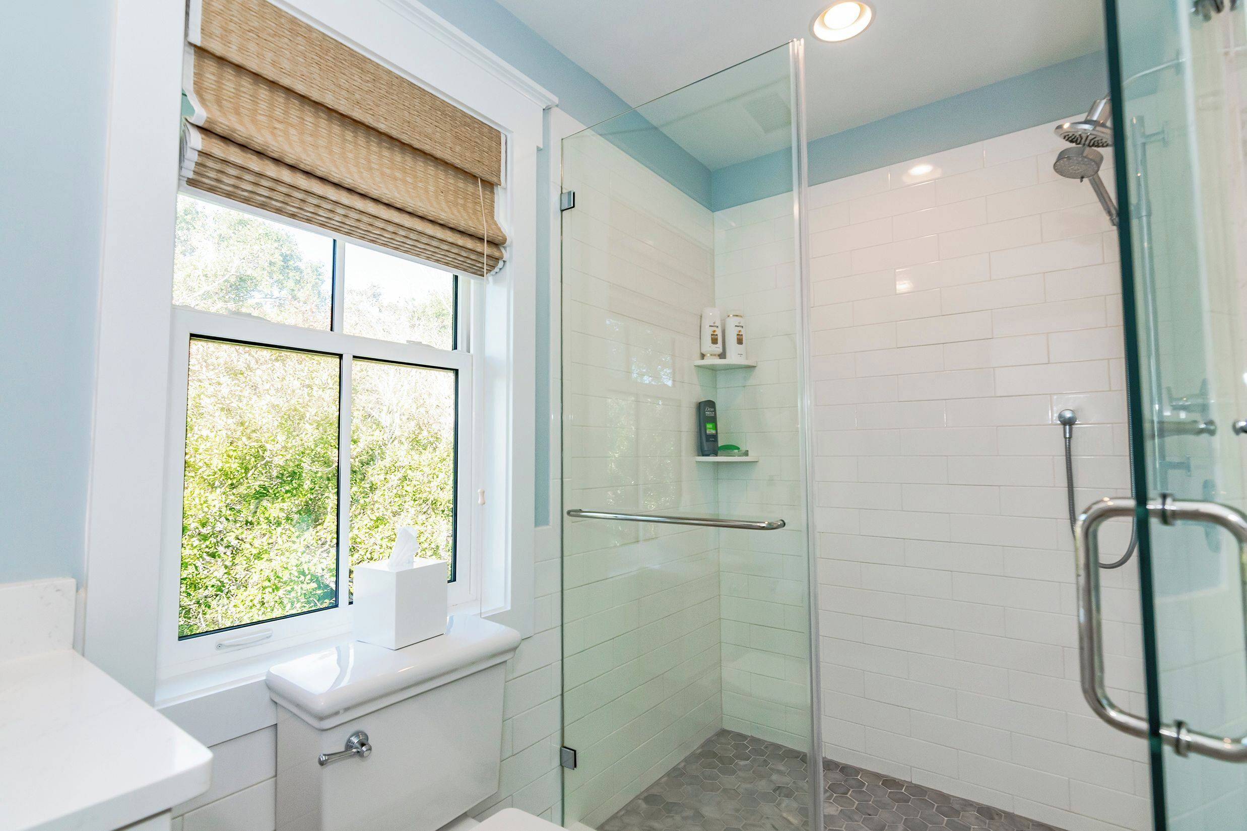 Une salle de bain moderne avec une douche à l’italienne en verre, une fenêtre donnant sur un espace verdoyant et un lavabo blanc.
