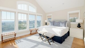 A bedroom with a large bed, two bedside tables, a nightstand, a window with a view, and a wooden bench. - Captain Tank's Ocean View with covered deck (Bald Head Island)