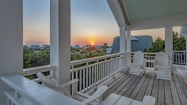 A covered outdoor deck with white Adirondack chairs and a view of the sunset over a residential area. - Captain Tank's Ocean View with covered deck (Bald Head Island)