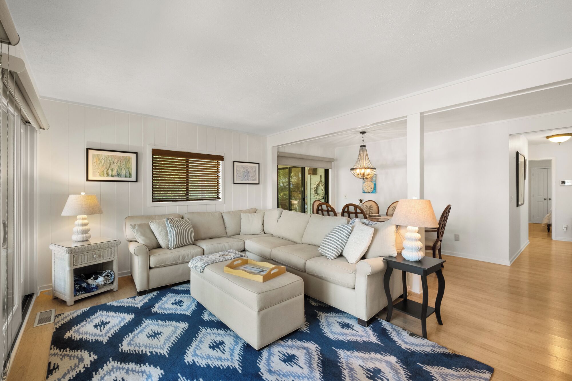 A living room with a sectional sofa, a coffee table, and two side tables with lamps. There are framed pictures on the walls and a window with blinds.
