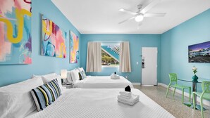 Une chambre d’hôtel avec deux lits, un ventilateur de plafond, une fenêtre avec des rideaux, une télévision et une petite table avec des chaises.