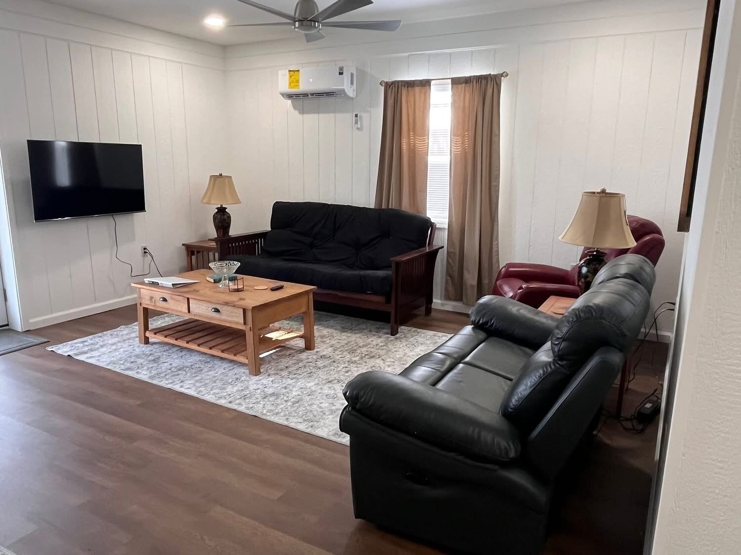 Una sala de estar con televisor de pantalla plana, una mesa de centro de madera, un sofá negro, un sillón rojo y una alfombra color beige.