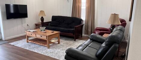 Una sala de estar con televisor de pantalla plana, una mesa de centro de madera, un sofá negro, un sillón rojo y una alfombra color beige.