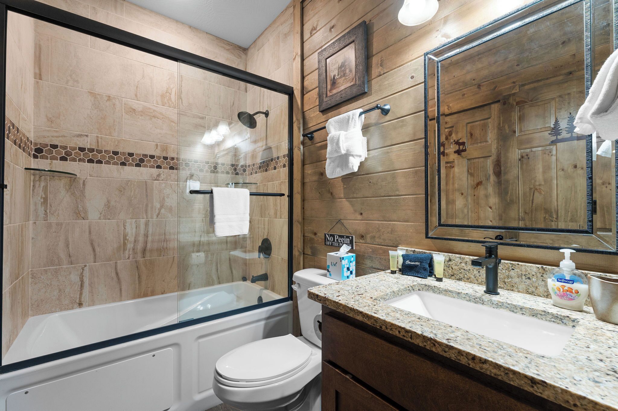 Un baño con ducha envuelta en cristal, inodoro blanco, lavabo con encimera de granito y un espejo grande.