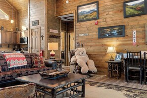 Interior rústico de una cabaña con paredes de madera, un sofá acogedor, un gran oso de peluche y cuadros en las paredes.