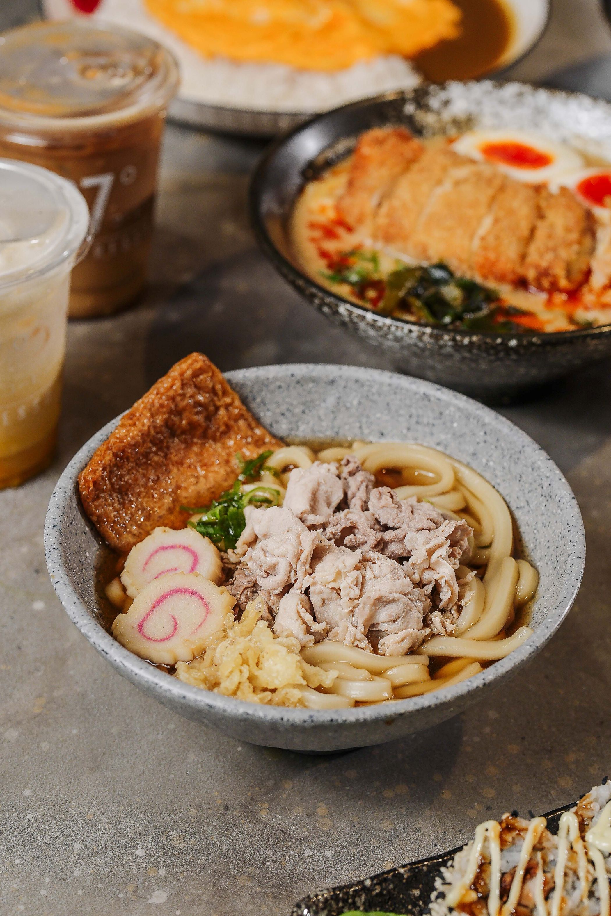 A bowl of ramen with various toppings, a cup of iced coffee, and a plate of food in the background.