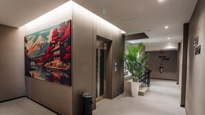 A hallway with an elevator, a trash bin, a potted plant, and a wall with a mural. - STAY KERTAPATI HOTEL (Palembang)