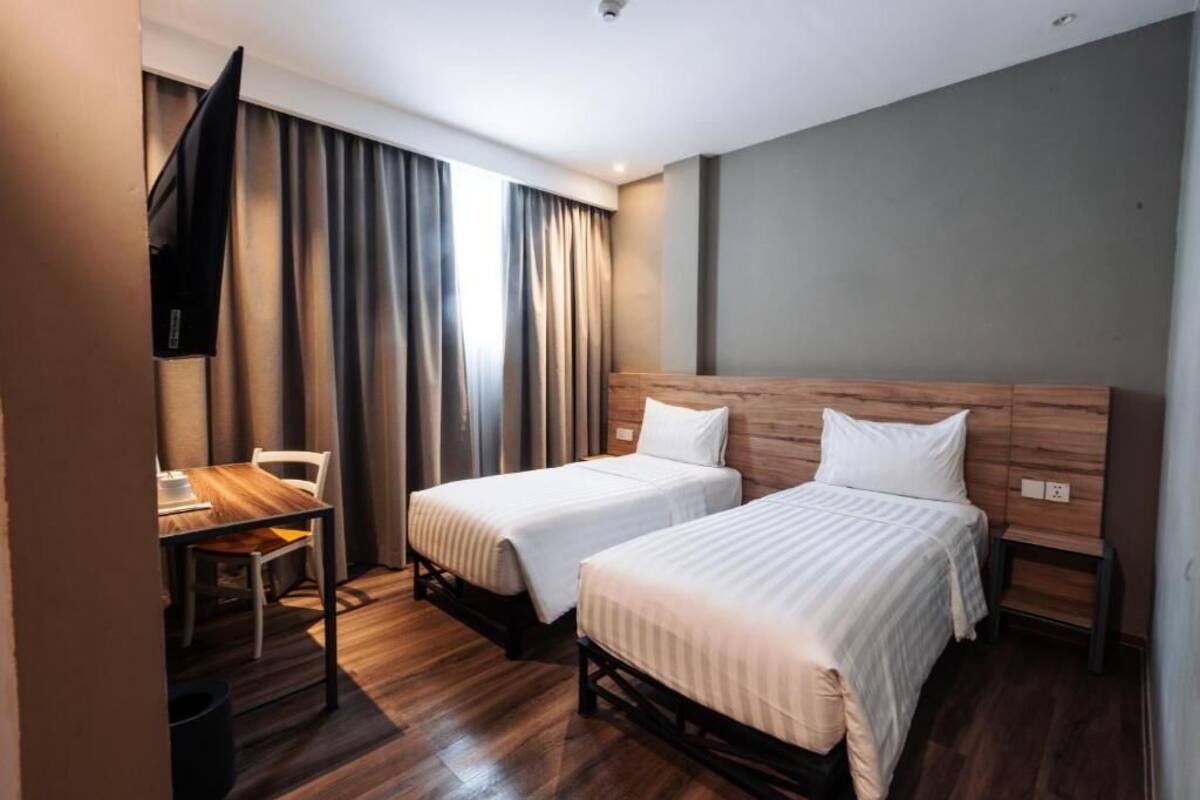 A hotel room with two single beds, a wooden headboard, a small table with a chair, and a TV mounted on the wall.