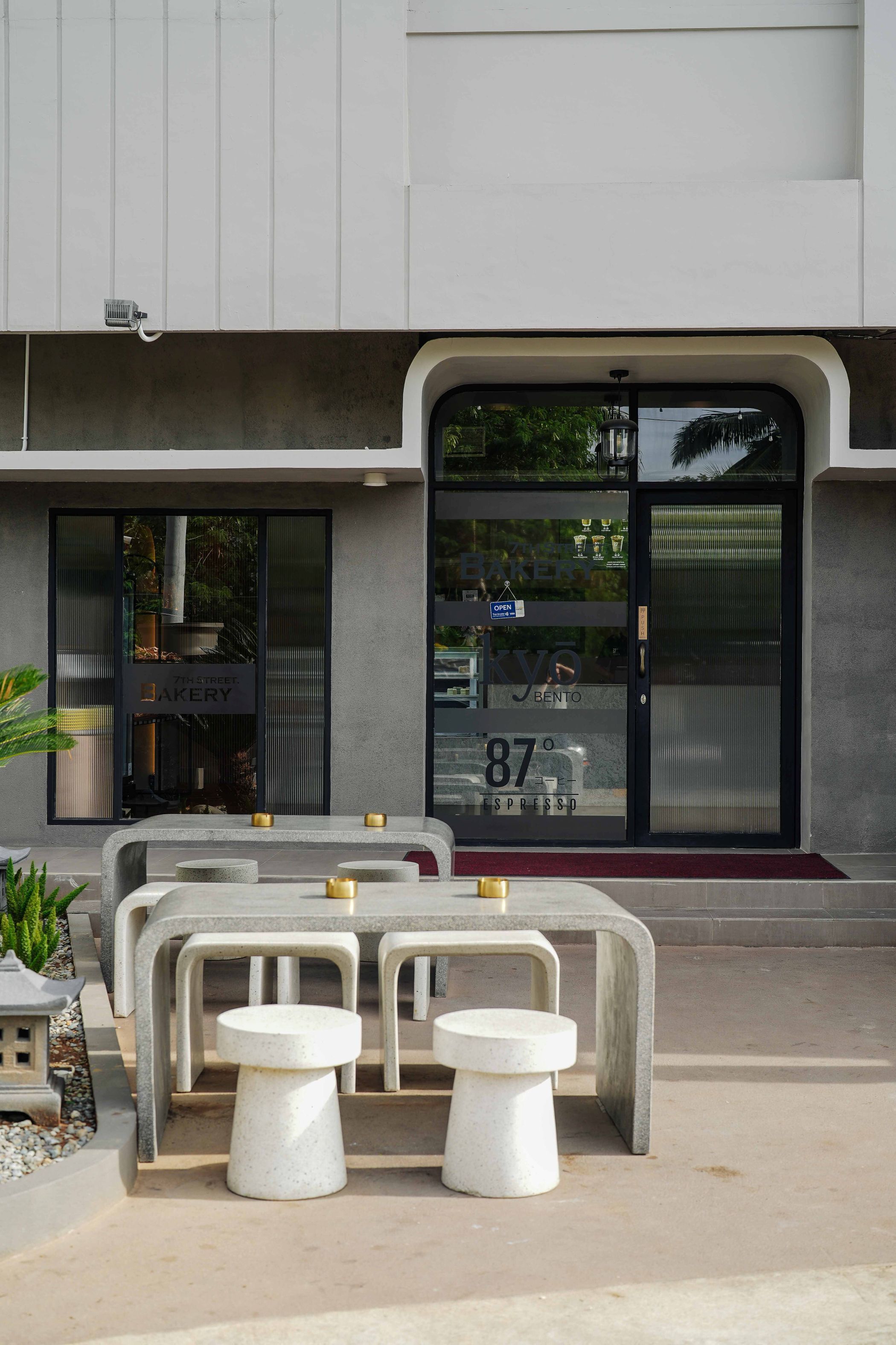 Un edificio moderno con escaparate, zona de estar exterior con mesas y taburetes de hormigón, y un amplio escaparate de vidrio con señalización.