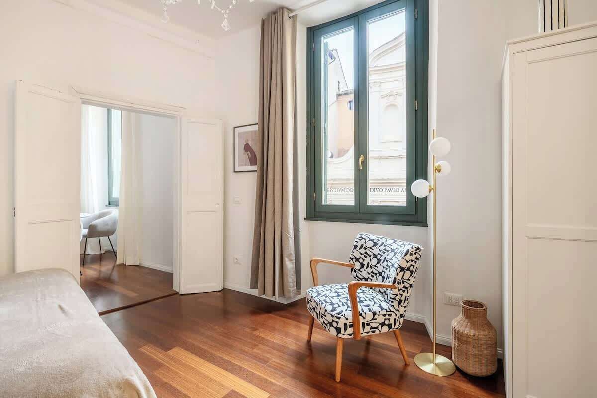 A room with a bed, a chair, a window with curtains, a floor lamp, and a vase.