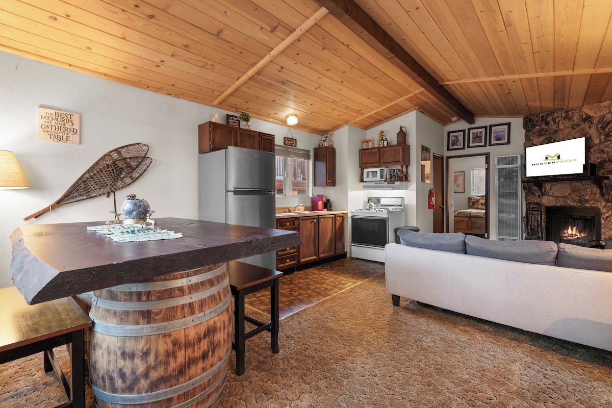 A rustic interior with a wooden ceiling, a kitchen area, a seating area with a fireplace, and a television mounted on a stone wall.