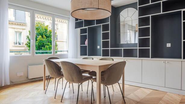 Een moderne keuken met een houten tafel en stoelen, inbouw zwarte planken en een groot raam met uitzicht op groen.