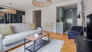 Een moderne woonkamer met een bank, salontafel en televisie. Een keuken is zichtbaar door een open doorgang.